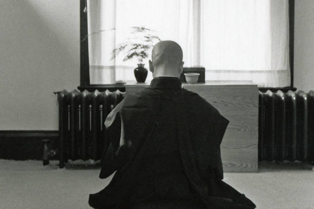 Black and white photograph of a robed figure seen from behind, sitting on a cushion facing a low altar beneath a tall curtained window