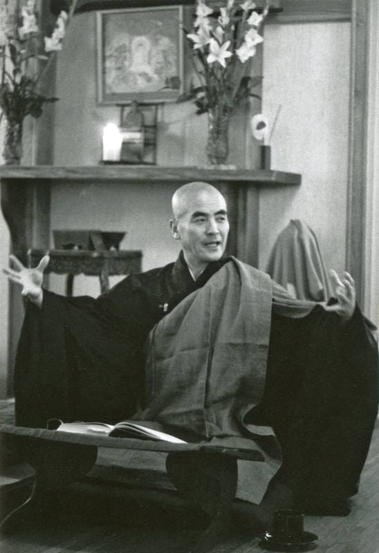 Black and white photograph of Katagiri Roshi in dark robes, seated behind a low lectern with an open book, smiling as he speaks with both hands raised; lilies and a candle on the altar behind him