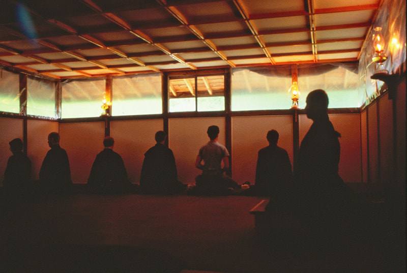 A row of people sitting zazen facing the walls of a wooden meditation hall, seen in silhouette against high windows and oil lamps; one person stands in the foreground at the right