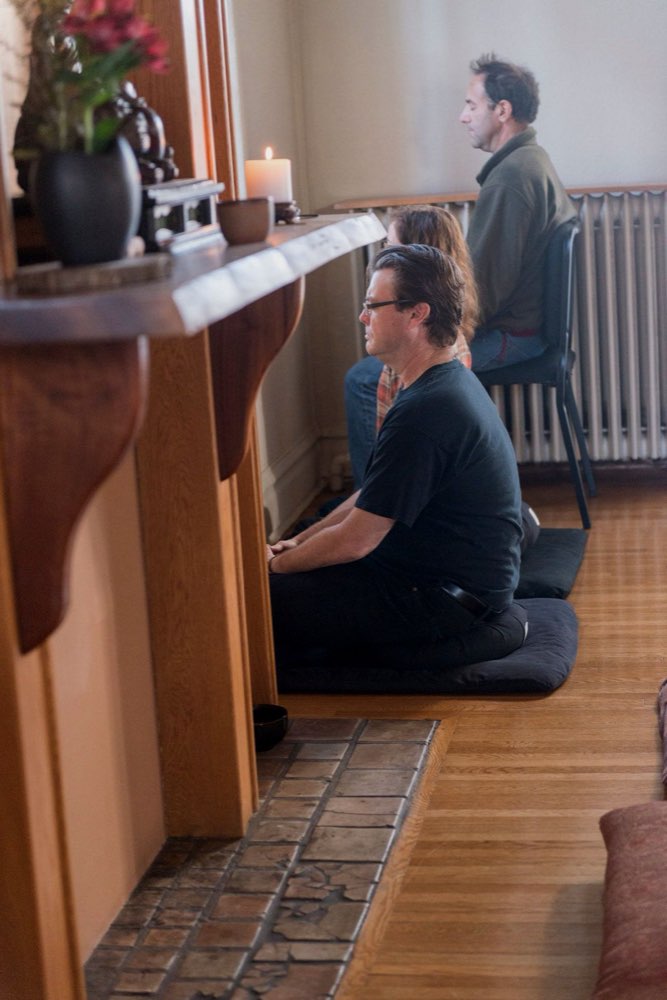 Two people sitting in meditation on black cushions, facing the wall of the meditation hall